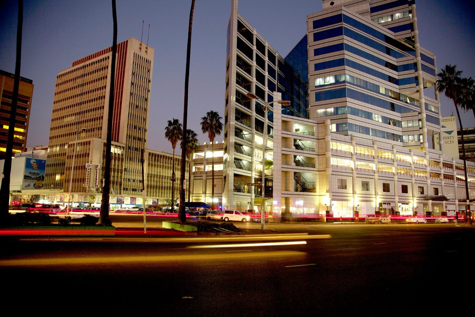 Harare's central business district at dusk: office towers lit above palm trees, with the light trails of passing traffic