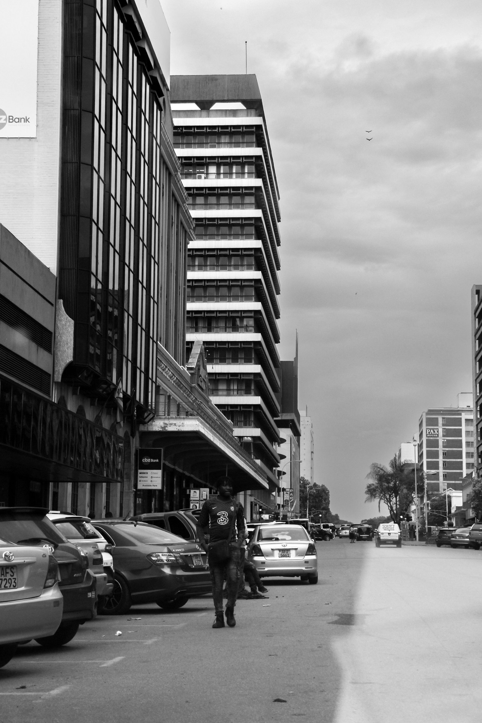 A city street in black and white, cars parked along the kerb beneath commercial frontages